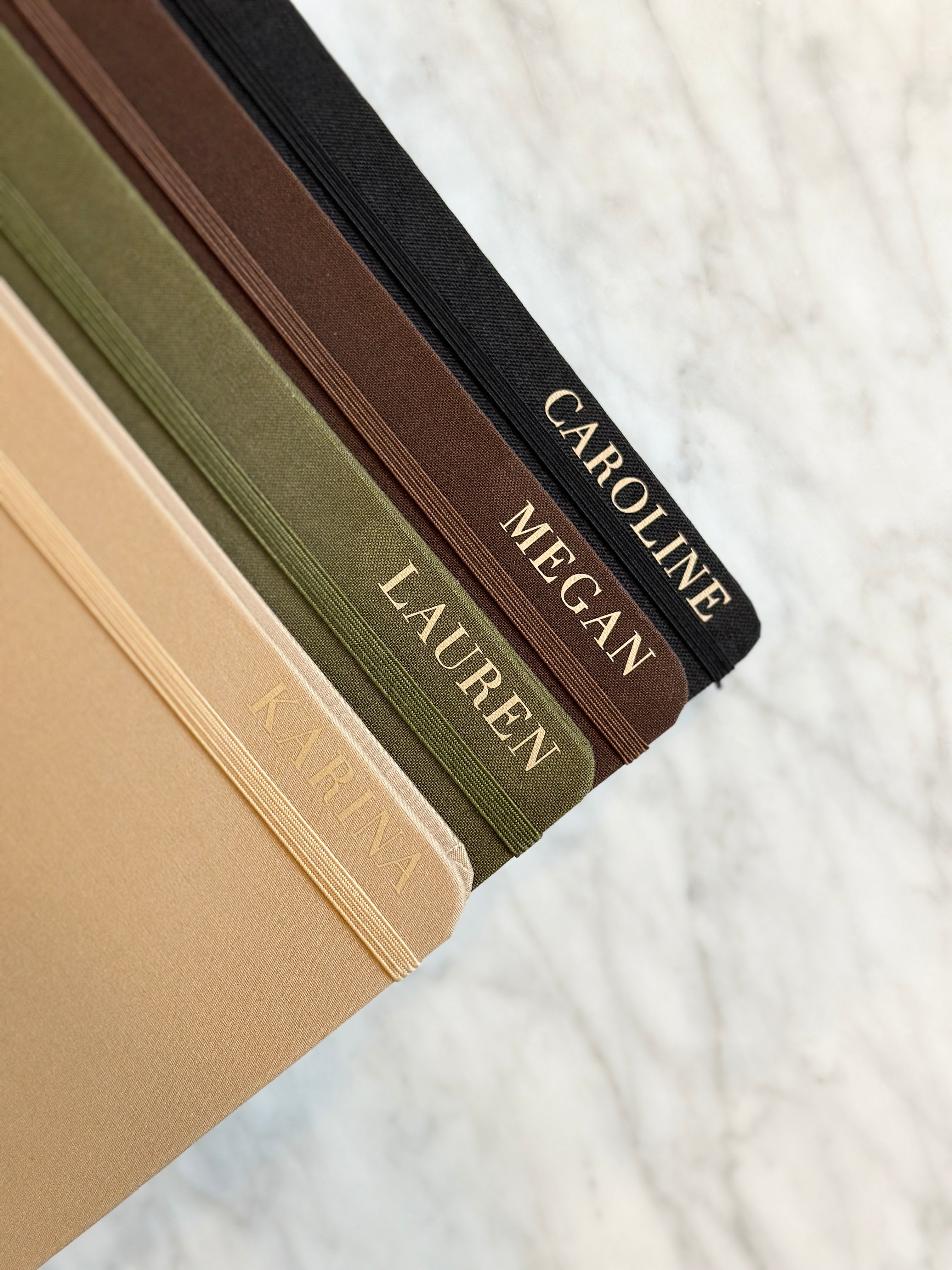 Luxury linen bullet journals in black, brown, olive, and beige with custom names on the front in gold.